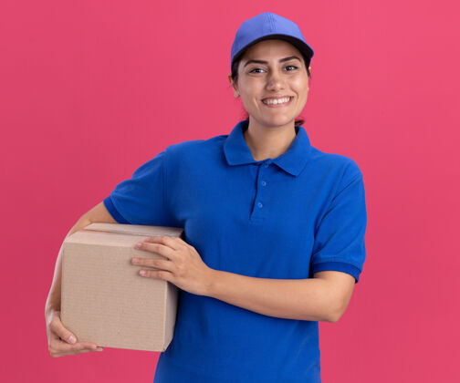 衣服带着微笑的年轻送货员穿着制服 粉色墙上有一个装帽子的盒子微笑递送抱着