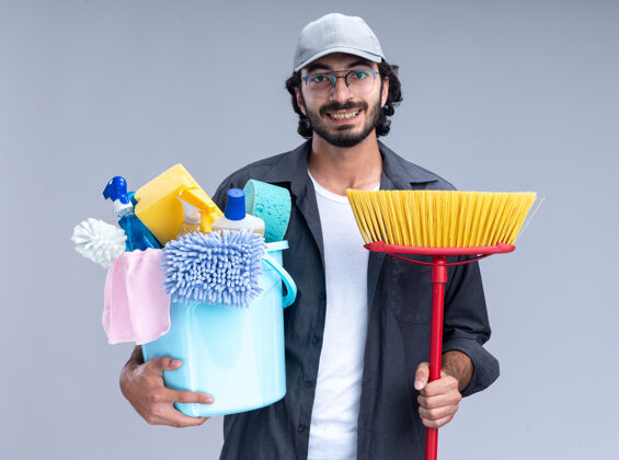 帅哥面带微笑的年轻帅气的清洁工穿着t恤 戴着帽子 手里拿着一桶清洁工具 把拖把隔离在白色的墙上表情拖把人