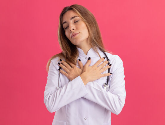 表情喜欢闭着眼睛的年轻女医生穿着带听诊器的医用长袍把手放在肩膀上隔离在粉红色的墙上人脸肩膀