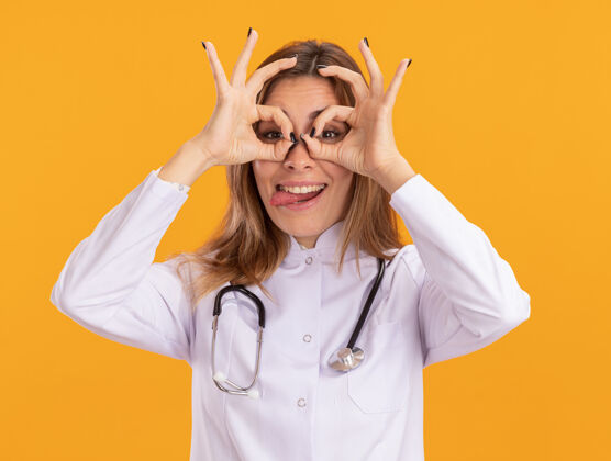 衣服快乐的年轻女医生穿着医用长袍 听诊器显示表情手势 舌头被隔离在黄色的墙上姿势女性快乐