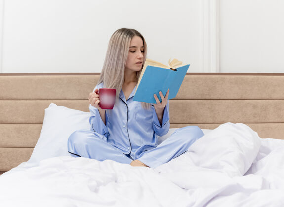 睡衣穿着蓝色睡衣的年轻漂亮女人坐在床上 在卧室里拿着一杯咖啡看书坐着杯子漂亮