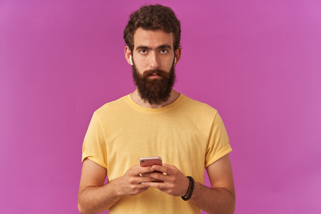 情感照片中细心帅气的留着胡子的年轻人手里拿着耳机 聚精会神地站在紫色的墙上模特聊天紫色