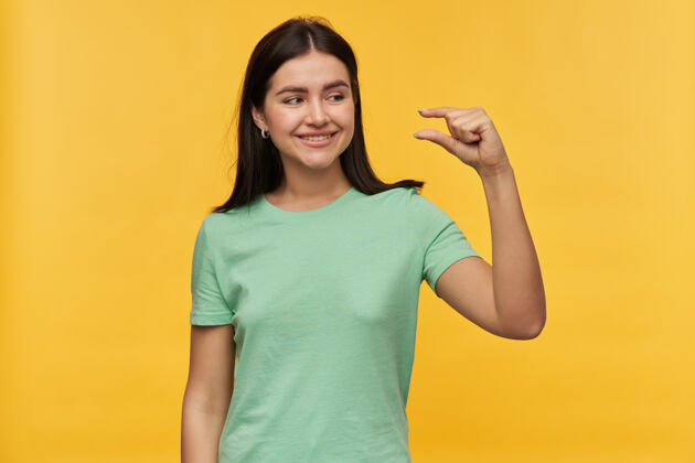 魅力微笑可爱的黑发年轻女子在薄荷tshirt站着 并显示由手指隔离在黄色的墙壁小尺寸年轻模特肖像