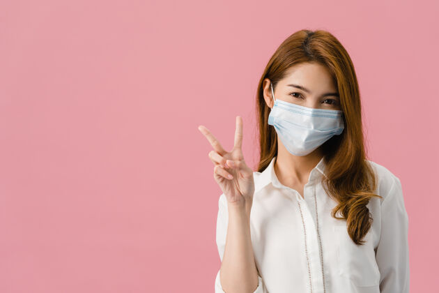 女性年轻的亚洲女孩戴着医用口罩显示和平的迹象 穿着休闲的衣服 看着隔离在粉红色背景上的相机鼓励卫生隔离流感