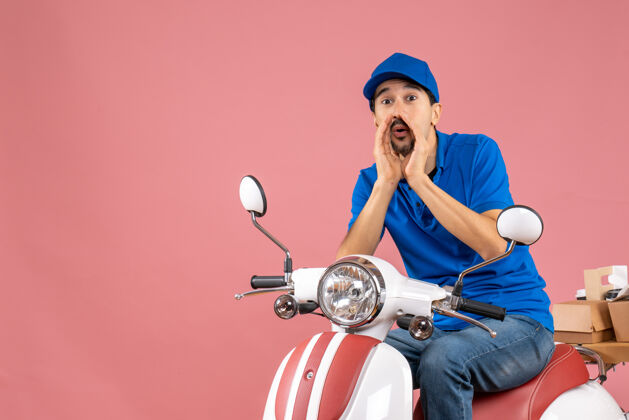 坐着一个戴着帽子的快递员坐在踏板车上 在柔和的桃色背景下给某人打电话踏板车前面摩托车