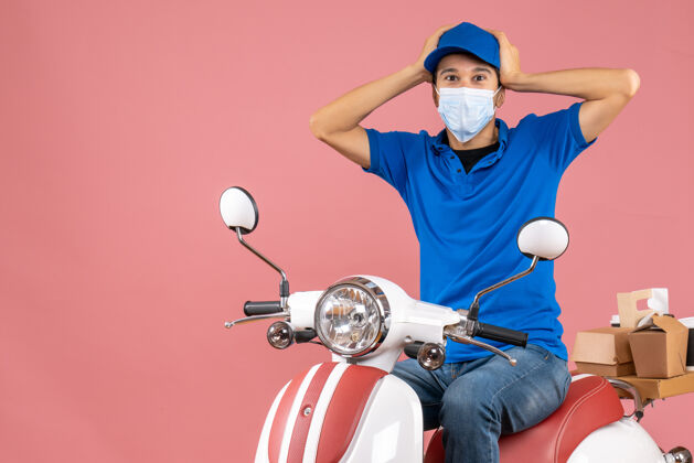 摩托车上图是一个戴着医学面具 戴着帽子 坐在粉彩桃色背景下的滑板车上 情绪混乱的送货员面具桃人
