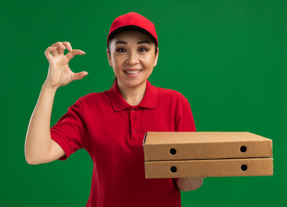 大小身穿红色制服 头戴鸭舌帽 手持披萨盒的年轻送货员站在绿色的墙上 微笑着摆出一副小巧玲珑的姿势拿着年轻手势