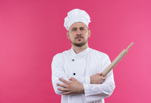 别针自信的年轻帅哥厨师身着厨师制服手拿擀面杖手放在手臂上隔离在粉红色的墙上帅气制服厨师
