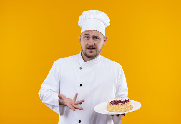 指着穿着厨师制服的年轻帅哥厨师手拿蛋糕盘 指着橘色墙上孤立的蛋糕厨师烹饪印象深刻