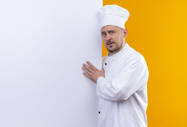 厨师穿着厨师制服的年轻帅哥厨师站在白色墙壁后面 用手指着隔离在橙色墙壁上的复制空间制服立场手