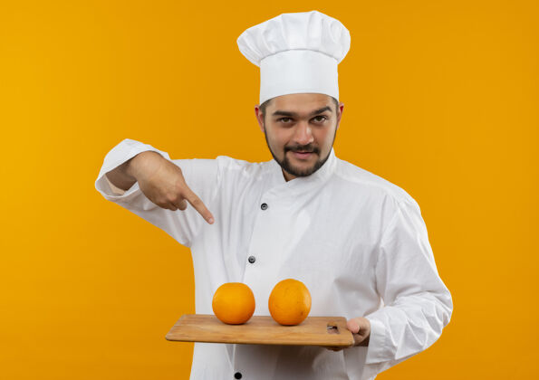 制服身着厨师制服的自信的年轻男厨师手拿着并指着隔离在橙色墙上的砧板 砧板上有橘子信心橙色男性
