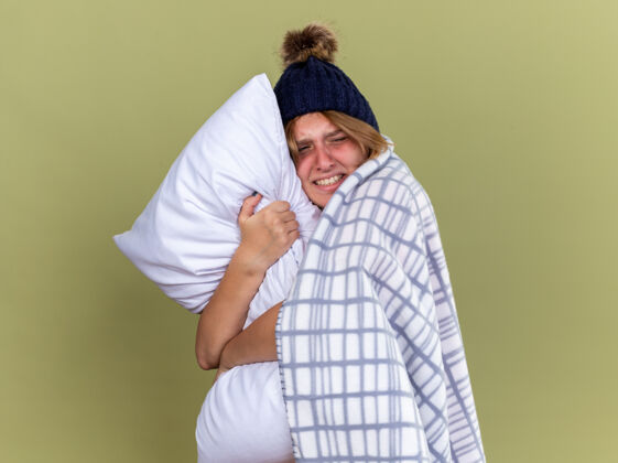抱着不健康的年轻女子裹着毛毯戴着帽子抱着枕头忍受着感冒和流感的折磨站在绿色的墙上感到恶心和烦恼站着痛苦烦恼