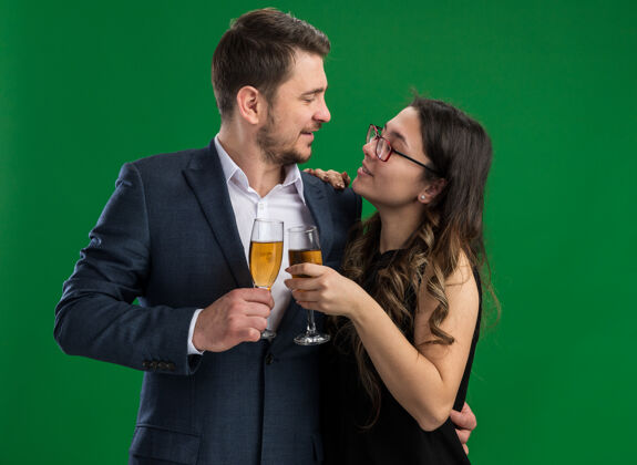 男人年轻漂亮的夫妇幸福的男人和微笑的女人 戴着香槟 拥抱着 看着彼此 相爱着 一起庆祝情人节 站在绿色的墙上一起眼镜拥抱