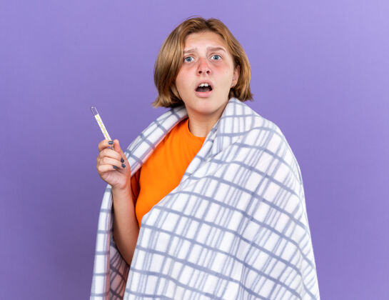 感觉不健康的年轻女子裹着温暖的毯子 感觉不舒服 患流感 发烧 用温度计测量体温 站在紫色的墙上 看起来很担心年轻发烧包裹