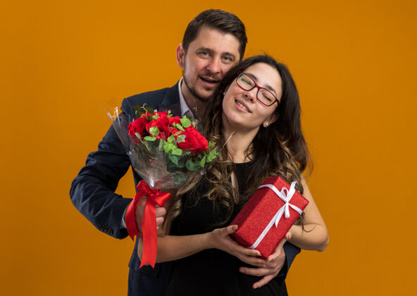 禮物幸福而美麗的一對 男人拿著玫瑰花束 女人拿著禮物 擁抱著幸福的愛情 在橙色的墻上慶祝情人節女人情侶花束