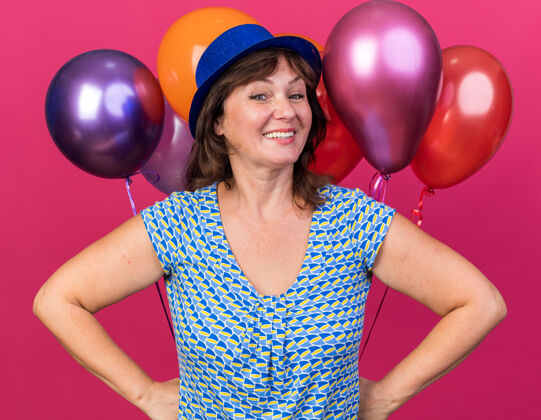女人快乐快乐的中年妇女 戴着派对帽 手持五颜六色的气球 站在粉色的墙上欢笑着庆祝生日派对举行微笑喜悦