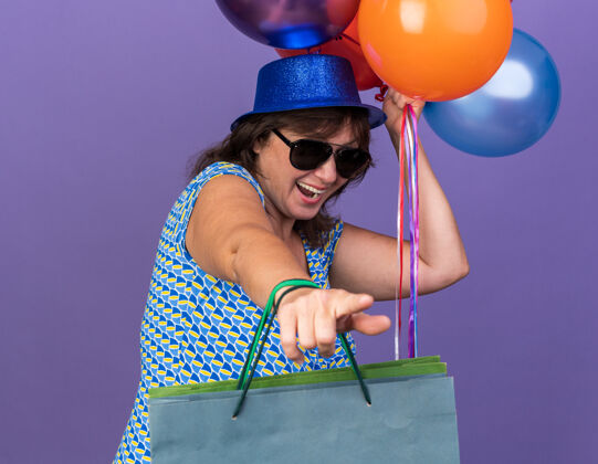 帽子快乐和兴奋的中年妇女戴着聚会帽和眼镜 手里拿着一堆五颜六色的气球和带着礼物的纸袋 站在紫色的墙上庆祝生日聚会气球五颜六色包