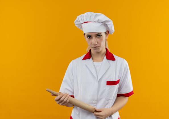 烹饪自信的年轻漂亮厨师穿着厨师制服拿着擀面杖孤立地放在橙色的墙上 留着复制空间漂亮别针自信