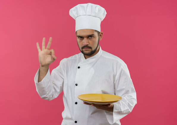 制服自信的年轻男性厨师 身着厨师制服 拿着盘子 在粉红色的墙上贴着“做得好”的牌子 并留有复印空间年轻自信好的