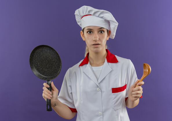 持有年轻漂亮的厨师穿着厨师制服 手拿煎锅和勺子 孤立地站在紫色的墙上锅制服薯条