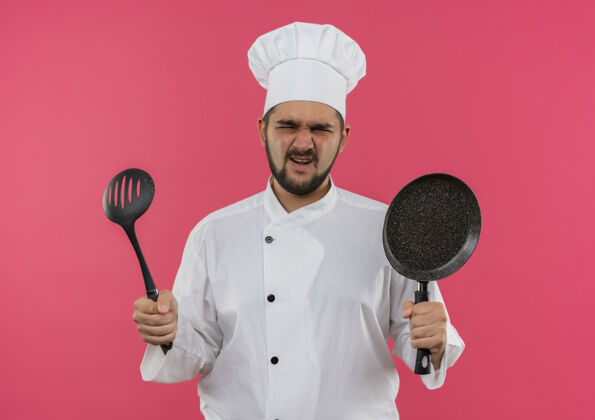 薯条恼火的年轻男厨师穿着厨师制服 手里拿着煎锅和一字汤匙 被隔离在粉红色的墙上厨师烹饪平底锅