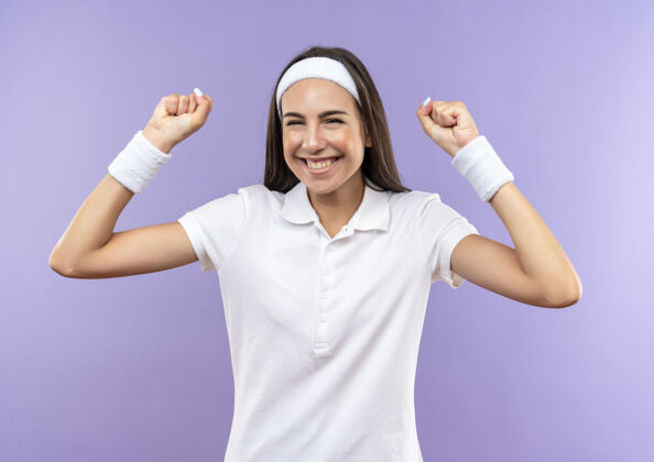 拳头快乐漂亮的运动女孩戴着头带和腕带举起拳头孤立在紫色的墙上戴上女孩头带