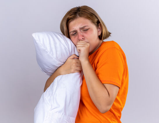 枕头不健康的年轻女子穿着橙色t恤 抱着枕头 感觉不舒服 患流感 拳头咳嗽 站在白墙上站痛苦女人