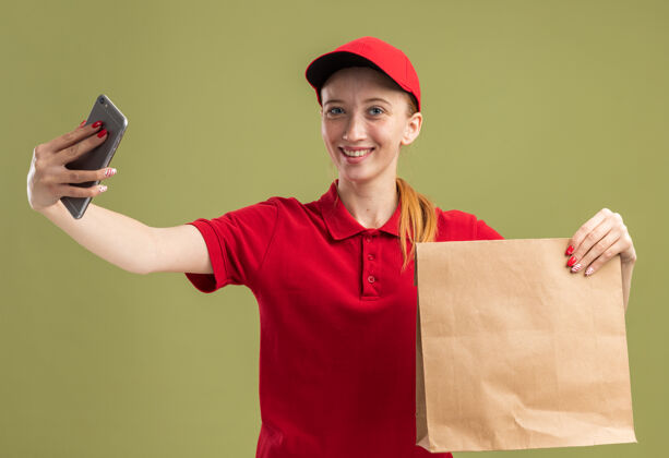 欢呼身穿红色制服 头戴鸭舌帽的年轻送货员拿着纸包 用智能手机自拍 在绿色的墙上欢快地笑着微笑抱着自拍