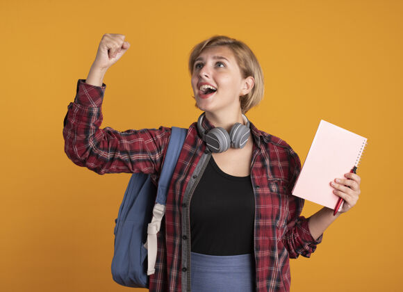 提高惊讶的年轻斯拉夫女学生戴着耳机 背着背包 举起拳头 拿着笔记本和笔 看着身边斯拉夫举行橙色