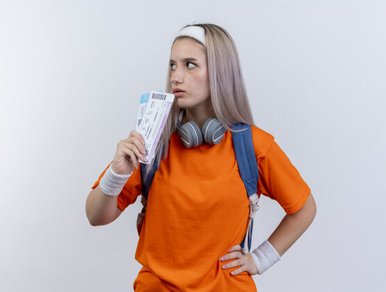头带自信的年轻白人运动女孩 戴着耳机 脖子上戴着背包 头上戴着头带和手腕 手里拿着机票 看着身边腕带年轻运动