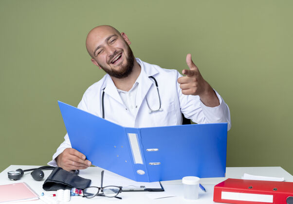 医生年轻的秃头男医生穿着医用长袍和听诊器坐在办公桌旁大笑工具笑医疗