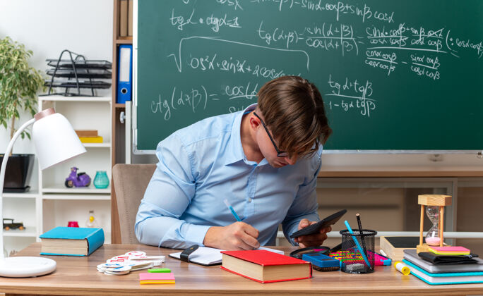 桌子年轻的男老师戴着眼镜 带着计算器备课 自信地坐在课桌旁 教室里的黑板前放着书和笔记坐着准备前面