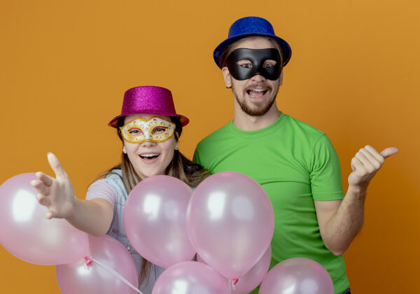 帅气快乐的年轻夫妇站在氦气球女孩在粉红色的帽子戴化装眼罩点向前与手帅哥在蓝色的帽子戴化装眼罩点在一边帽子眼睛侧线