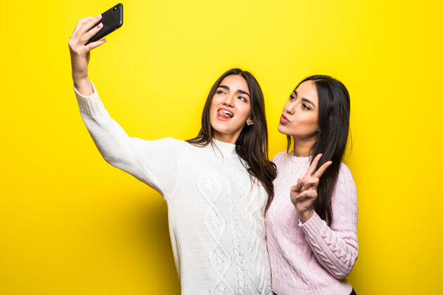 电话两个穿着毛衣的开朗女孩站在黄色墙壁上自拍的画像毛衣制造时尚