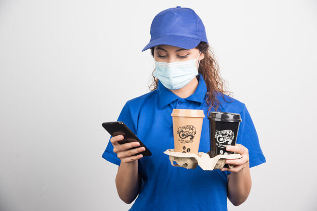女性穿着蓝色制服 戴着医用口罩的女人看着电话 手里拿着两杯白咖啡优惠女士手机