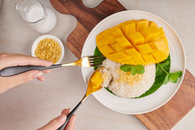 持有熟透的芒果和糯米 在石头表面的盘子里放上椰奶 夏天的泰国甜点女人用叉子和勺子吃芒果和糯米俯瞰图泰国木头女孩