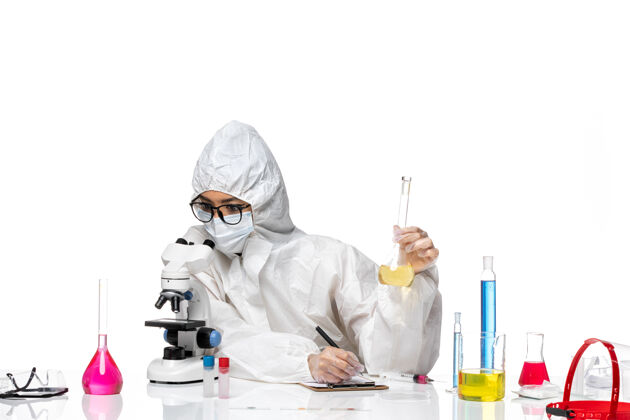 套装正面图身着特殊防护服的年轻女化学家拿着装有溶液的烧瓶 用显微镜观察白色背景的实验室冠状病毒化学病毒防护医生医学