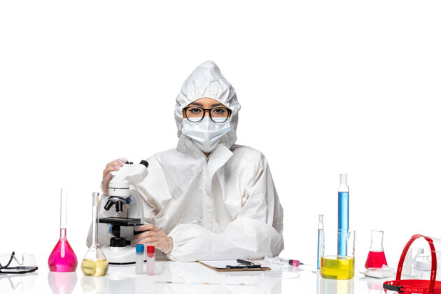 使用正面图身着特殊防护服的年轻女化学家在白色办公桌上使用显微镜病毒实验室共价健康化学面具显微镜化学