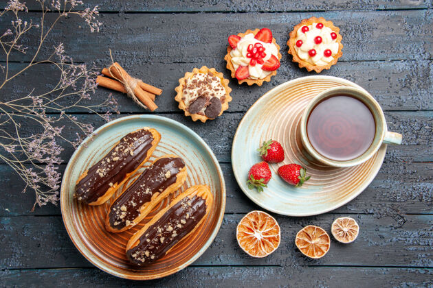 深色俯瞰图：椭圆形盘子上放着巧克力 茶托上放着一杯茶和草莓 黑木桌上放着肉桂和柠檬干木头扣子茶