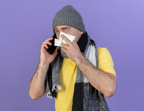 电话焦急的年轻金发病斯拉夫男子戴冬季帽子和围巾擦鼻子讲电话斯拉夫湿巾鼻子