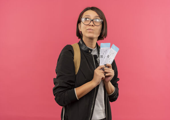 衣服体贴的年轻女学生戴着眼镜背着包拿着机票看着隔离在粉红色墙上的一面粉色表情脸