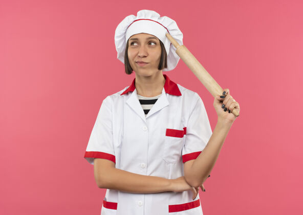表情体贴的年轻女厨师身着厨师制服 手持擀面杖 把手放在胳膊肘下 看着粉色墙壁上孤立的一面滚动手人