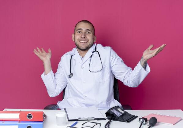 粉色面带微笑的年轻男医生 穿着医用长袍 听诊器 坐在办公桌旁 手里拿着工作工具 两手空空的 被隔离在粉红色的墙上男性微笑年轻