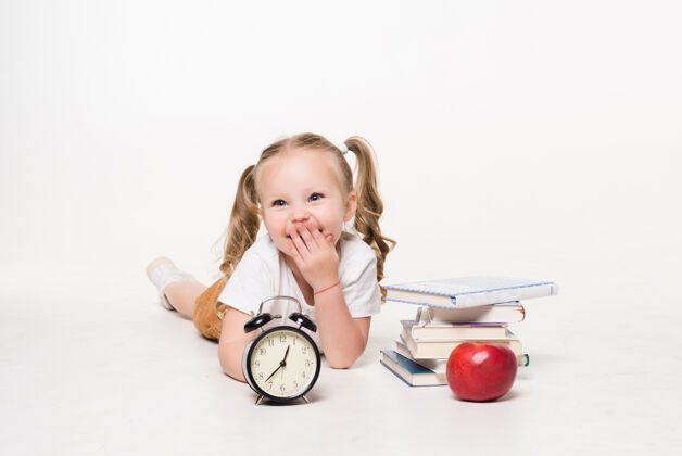 孩子教育和学校理念满脸笑容的小女孩拿着书钟和苹果躺在地上女学生时钟红苹果