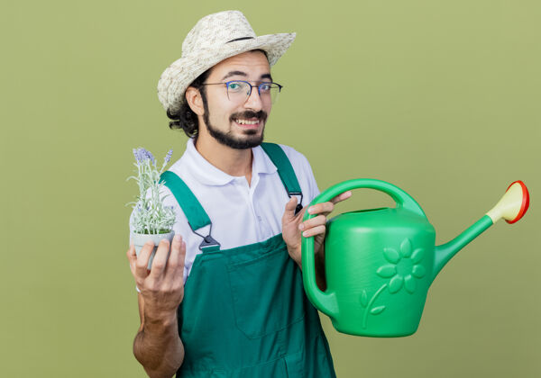 人年轻的留着胡须的园丁 穿着连体衣 戴着帽子 拿着水罐和盆栽植物 站在浅绿色的墙上 面带微笑地看着前方表情微笑连身衣