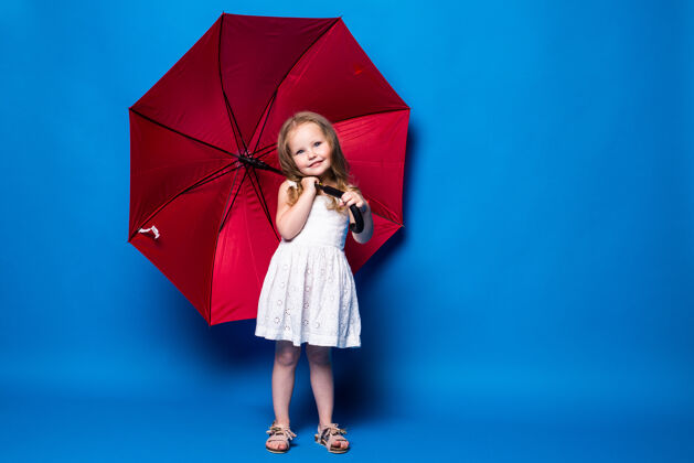伞快乐的小女孩拿着红伞在蓝色的墙上摆姿势可爱白裙子漂亮