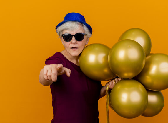 姿势戴着派对帽 戴着太阳眼镜的自信的老妇人站在那个里 氦气球指着橙色墙壁上孤立的前方站气球女人