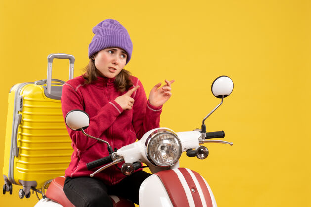 年轻女子前视图：骑着轻便摩托车戴着紫色帽子的年轻女子指着后面帽子指尖快乐