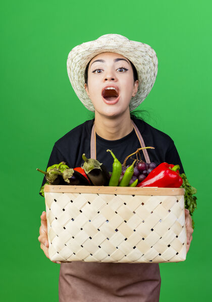 衣服年轻漂亮的女园丁 围着围裙 戴着帽子 手里拿着装满蔬菜的篮子 站在绿色的墙上 满脸幸福地喊叫着喊叫蔬菜女人