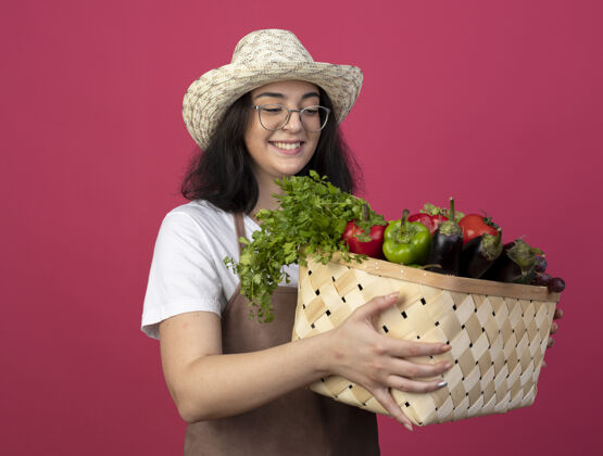园艺微笑着的年轻黑发女园丁 戴着眼镜 穿着制服 戴着园艺帽 站在粉红色的墙上看着孤立的菜篮子蔬菜市民姿势
